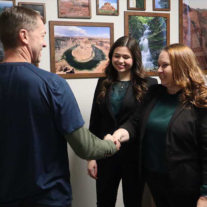 A man in a blue shirt shakes hands with a woman in a black blazer, while another woman looks on, against a backdrop of nature photos.