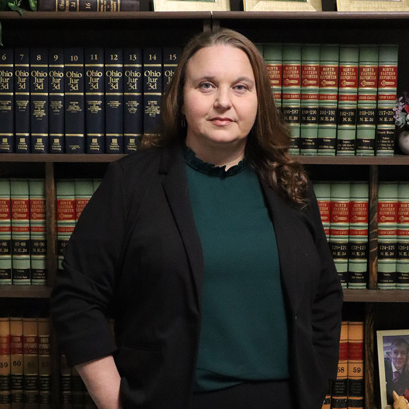 A woman in professional attire stands before a bookshelf filled with law books and framed certificates, conveying a scholarly atmosphere.