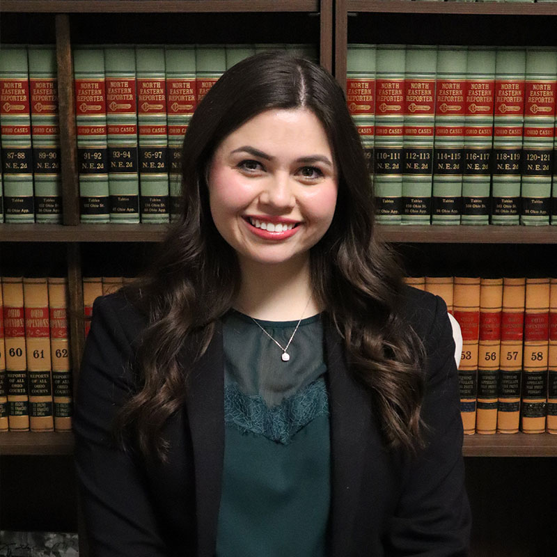 A woman with long, wavy hair sits in front of a bookshelf filled with law books, dressed in a dark blazer and teal top.