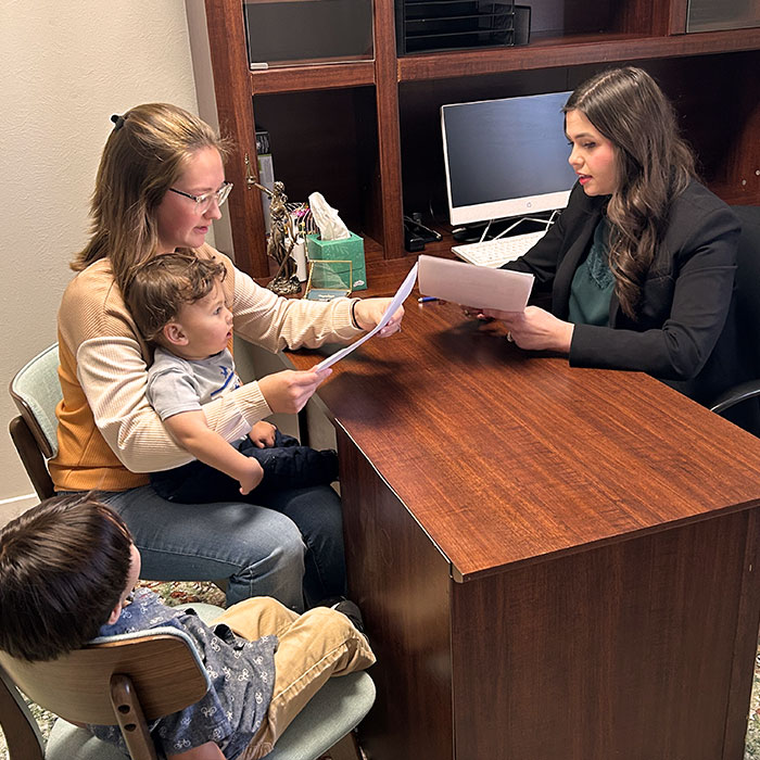 A woman seated at a desk reviews documents with another woman holding a toddler, while a young child watches from a nearby chair.
